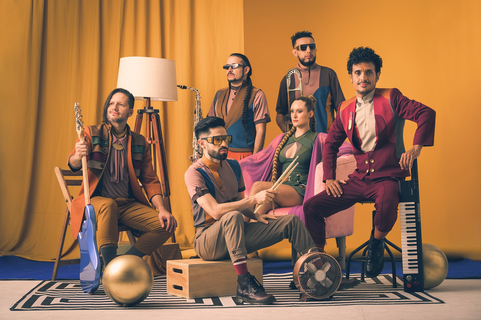 Photo du groupe Puerto Candelaria. Les six membres du groupe sont présents dans une salle aux teintes orangées. Ils sont habillés avec des vêtements colorés, aux styles modernes et créatifs. Chacun tient son instrument de musique entre ses mains.